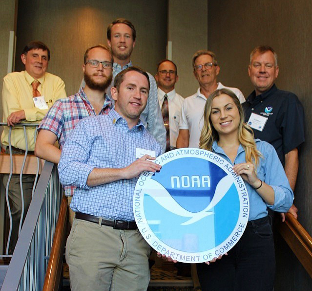 Delegation of eight NOAA employees standing on a staircase holding a large plaque with the NOAA emblem in front of them at the Eighth Biennial Southwest Aviation Weather Safety Workshop in Tempe, Arizona