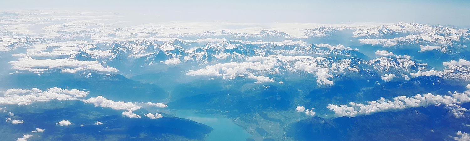 Aerial view of a mountain range with snow-capped peaks, clouds, and a lake nestled among the mountains.